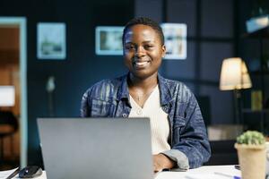 Preto mulher trabalhando às dela computador portátil dentro uma moderno apartamento, estudando conectados e pesquisando em formação para dela projeto. sorridente africano americano blogueiro usando pessoal computador enquanto olhando às Câmera. foto