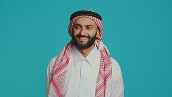 muçulmano adulto dentro tradicional árabe vestir, sorridente e em pé com confiança enquanto vestindo xadrez padronizar cachecol e branco vestido, chocando islâmico cultura. jovem sério feliz árabe cara. foto