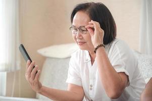 idosa mulher asiática sentada no sofá está fazendo uma videochamada no smartphone. a outra mão agarra os copos.