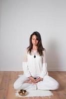 linda mulher adulta vestida de branco durante uma sessão de meditação. sentado de pernas cruzadas pose de ioga. tigela de canto de copa. foto