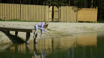 jovem mulher é sentado em Beira do cais sozinho. estoque imagens de vídeo. lindo jovem mulher lança pedras para dentro água em de madeira cais. jovem mulher é sentado em cais e jogando pedras em ensolarado verão dia foto