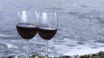 romântico dia com óculos do vermelho vinho em pé em a de praia em uma ensolarado dia. conceito. dois transparente óculos com vermelho vinho em uma seixo de praia com ondas e mar em a fundo. foto