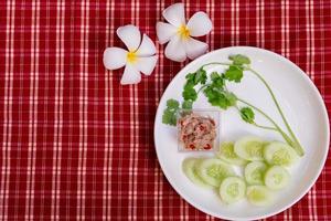 A salada de atum yum é pequena em um prato branco, guarnecida com melões e coentros verdes frescos. fundo de tecido xadrez vermelho e branco. estilo vintage. decorado com flores de frangipani. foto