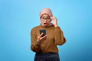 jovem asiática surpresa olhando para a tela do celular com a boca aberta sobre fundo azul foto