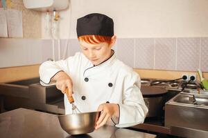 ruivo Garoto dentro chef traje cozinheiros panquecas dentro cozinha, amassa massa, assistente faz café da manhã. tocam com batedor foto