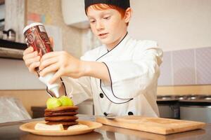 criança Garoto dentro chef traje prepara chocolate panquecas dentro cozinha dentro cafeteria. derrama chocolate foto