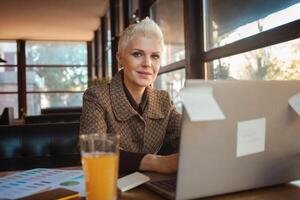 Senior mulher do 50. anos é lindo e à moda, senta dentro cafeteria, trabalho com computador portátil, freelance designer. parece ótimo, feliz vida, ativo estilo de vida. parece para dentro Câmera. foto