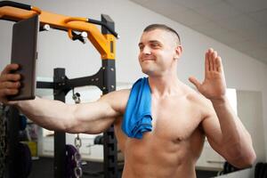 muscular bonito homem treinador é fotografado depois de treinamento, em repouso. telefone, tábua, vídeo chamar, moderno tecnologia, comunicação interior. ginásio. foto