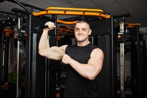 jovem bonito treinador dentro uma Preto uniforme contra fundo do uma simulador dentro academia. muscular Atlético corpo do uma fisiculturista, treinamento, Individual Esportes e peso perda retrato. músculos em a braço foto