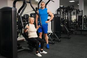 uma bonito jovem treinador homem ajuda a velho mulher trem Esportes dentro academia. feliz pessoas sorriso, apreciar vida. ativo, saudável estilo de vida, Senior. olhando para dentro a Câmera foto