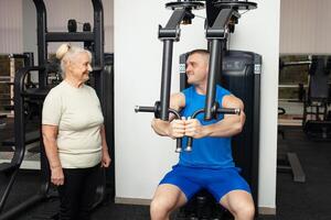 bonito jovem treinador homem mostra Senior mulher quão para devidamente executar exercícios em a simulador dentro a academia. feliz pessoas sorriso, aproveitar, feliz. ativo Esportes saudável estilo de vida interior. retrato foto