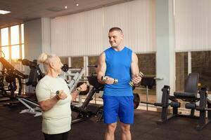 a velho mulher e uma jovem masculino treinador Faz exercícios dentro academia. elas mexe seus músculos com halteres, sorriso, estão feliz. ativo, saudável, desportivo estilo de vida, Senior conceito interior. luz solar foto