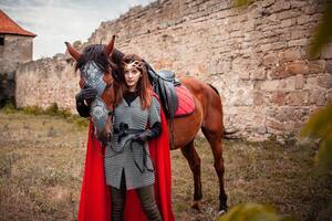 lindo Princesa com vermelho capa carrinhos Próximo para a cavalo contra a pano de fundo do uma torre e uma pedra parede foto