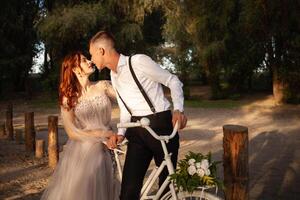 uma lindo casal dentro a madeiras em uma branco bicicleta com uma ramalhete do flores Casamento andar, uma mulher dentro uma cinzento vestir, uma homem dentro uma terno e suspensórios olhando às cada de outros foto