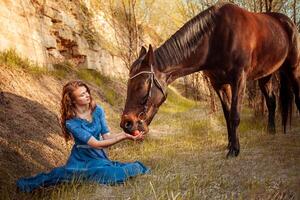 lindo menina dentro uma azul vestir feeds uma cavalo com a maçã foto