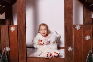 uma pequeno menina com brinquedos dentro uma de madeira casa. foto estúdio com Natal decorações. uma feliz criança tocam com brinquedos