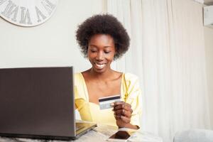 lindo africano americano Preto mulher usa uma banco cartão para compras on-line. Entrega do bens para casa. computador portátil em mesa foto