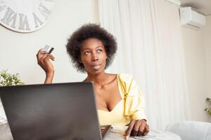 lindo africano americano Preto mulher usa uma banco cartão para compras on-line. Entrega do bens para casa. computador portátil em mesa. acha, sonhos, escolhe o que Comprar foto