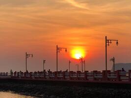 paisagem urbana abraçado de a caloroso brilho do pôr do sol com recortado torres, industrial estruturas, e uma sereno cais de a água, Como a céu tintas uma paleta do laranja matizes dentro a cedo tarde luz foto