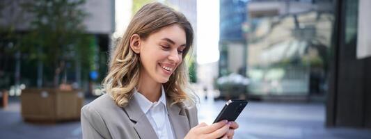 retrato do sorridente empresária bate-papo dentro aplicativo, olhando às Smartphone com satisfeito face, ordem Entrega para escritório, lendo mensagem em Móvel telefone foto