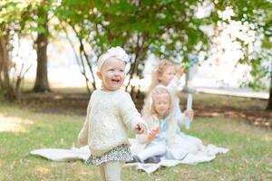 feliz pequeno menina dentro branco collants e bandanas em uma piquenique com dela irmãs. a bebê é Aprendendo para andar. foto