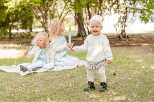 feliz pequeno menina dentro branco collants e bandanas em uma piquenique com dela irmãs. a bebê é Aprendendo para andar. foto