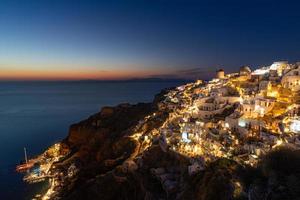 linda ilha do pôr do sol de santorini, grécia, bela vila caiada de branco, igreja e moinho de vento de oia durante o pôr do sol, luzes das ruas, férias de verão românticas, destino de viagem de casal famoso em lua de mel foto