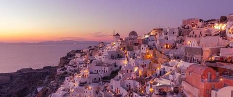 linda ilha do pôr do sol de santorini, grécia, bela vila caiada de branco oia com moinho de vento da igreja durante o pôr do sol, luzes das ruas, férias românticas de verão, destino de viagem de casal famoso em lua de mel foto