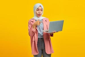 retrato de uma jovem asiática animada segurando um laptop e apontando isolado sobre um fundo amarelo foto
