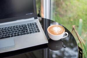 computador portátil com café com leite quente café dentro branco copo em escrivaninha foto