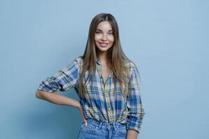 confiante jovem mulher dentro casual xadrez camisa posando com mão em quadril, grandes cabelo, sorridente, contra uma azul fundo foto