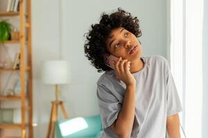 africano americano menina falando em Smartphone às casa interior. jovem mulher com célula telefone conversando com amigos. sorridente adolescente menina fazer respondendo ligar de celular. senhora tendo conversação de Móvel foto