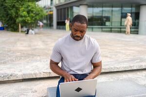 africano americano homem trabalhador autonomo usando computador portátil digitando conversando em urbano rua dentro cidade. cara tendo virtual encontro conectados bate-papo conferência. empreendedor programador trabalhando em computador portátil ao ar livre. controlo remoto trabalhos foto