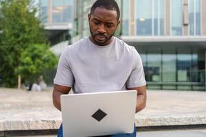 africano americano homem trabalhador autonomo usando computador portátil digitando conversando em urbano rua dentro cidade. cara tendo virtual encontro conectados bate-papo conferência. empreendedor programador trabalhando em computador portátil ao ar livre. controlo remoto trabalhos foto