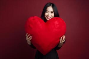 ai gerado sorridente ásia mulher segurando grande vermelho coração travesseiro em Sombrio vermelho fundo, neural rede gerado fotorrealista imagem foto