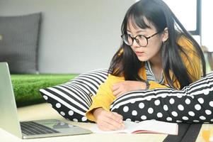 estudante adolescente dorme em travesseiros, lê o livro e usa o laptop. Ela faz anotações para se preparar para o exame. foto
