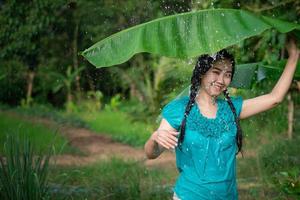 retrato de uma jovem asiática com cabelo preto segurando uma folha de bananeira na chuva no fundo do jardim verde