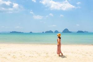 jovem, com um vestido branco, em pé na praia, olhando para o mar azul em um dia ensolarado durante as férias