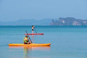 pai sênior asiático jogando stand up paddleboard ou sup com filha no mar azul nas férias de verão. conceito de família unida