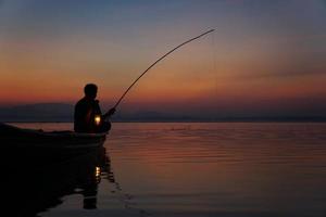 ao lado do lago, pescador asiático sentado em um barco e usando uma vara de pescar para pescar ao nascer do sol foto