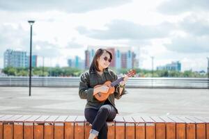 jogando ukulele do jovem lindo ásia mulher vestindo Jaqueta e Preto jeans posando ao ar livre foto