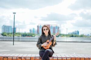 jogando ukulele do jovem lindo ásia mulher vestindo Jaqueta e Preto jeans posando ao ar livre foto