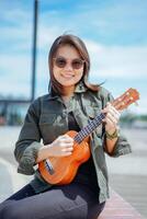 jogando ukulele do jovem lindo ásia mulher vestindo Jaqueta e Preto jeans posando ao ar livre foto