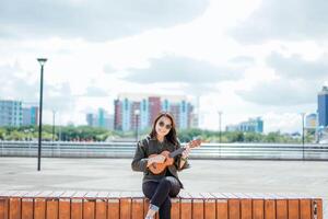 jogando ukulele do jovem lindo ásia mulher vestindo Jaqueta e Preto jeans posando ao ar livre foto