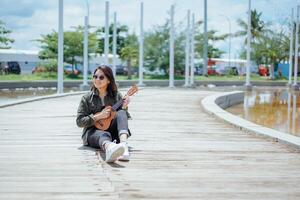 jogando ukulele do jovem lindo ásia mulher vestindo Jaqueta e Preto jeans posando ao ar livre foto