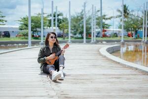 jogando ukulele do jovem lindo ásia mulher vestindo Jaqueta e Preto jeans posando ao ar livre foto