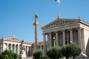 edifício neoclássico da academia de atenas com estátuas, bandeira e céu azul foto