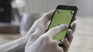 fechar acima do mãos usando Smartphone com verde tela, deslizante e tocando em borrado cozinha mesa fundo. estoque imagens de vídeo. homem mãos segurando Móvel telefone com croma chave. foto