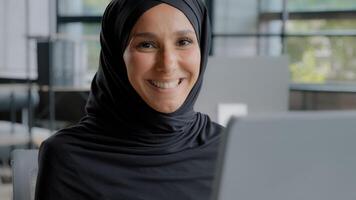 fechar-se positivo jovem mulher escritório trabalhador profissional Gerente trabalhando em computador bem sucedido atraente árabe empresária dentro hijab sentado às local de trabalho olhando às Câmera sorridente desfrutando fluxo de trabalho foto