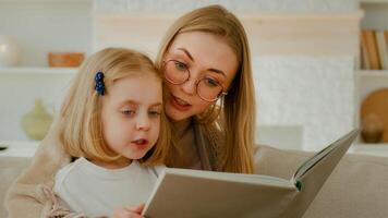 caucasiano mãe dentro Óculos com pequeno pequeno fofa filha pré escola criança menina bebê lendo livro dentro vivo quarto às casa mãe com criança ler literatura às sofá sofá elementar Aprendendo Educação foto